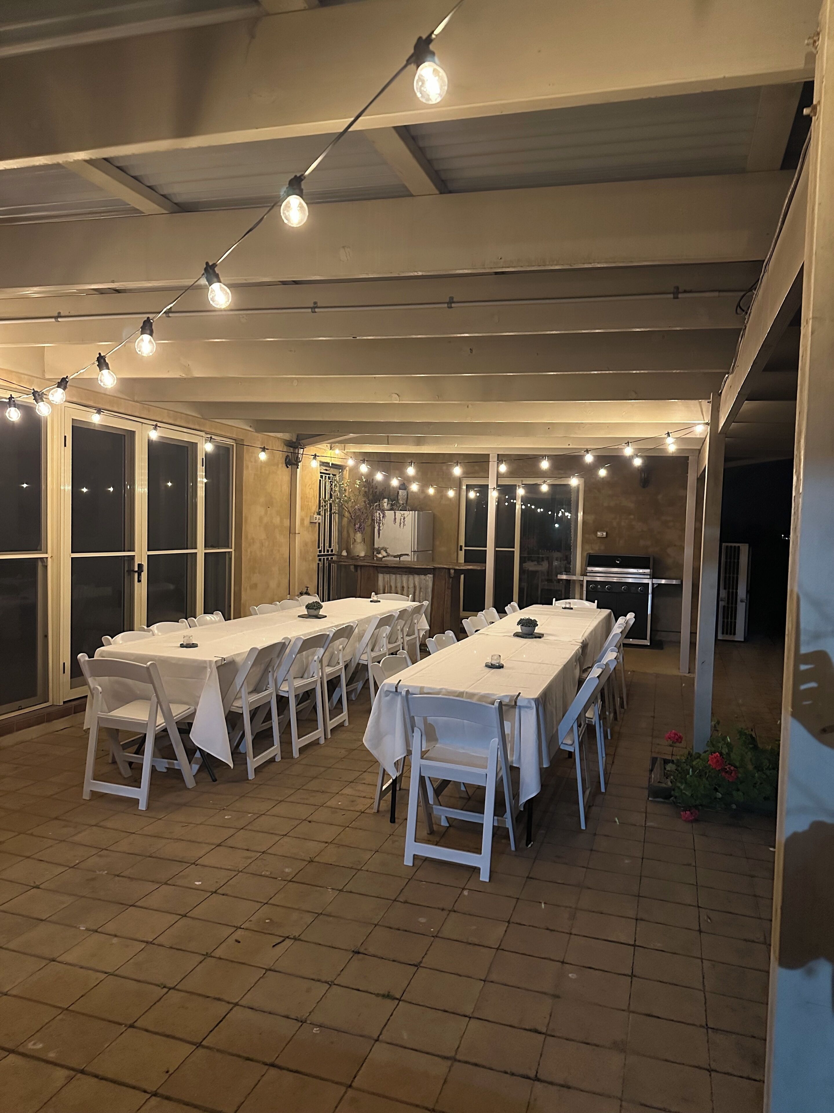 Un espacio comedor al aire libre techado con varias mesas y sillas, luces colgantes y un refrigerador.