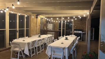 Un espacio comedor al aire libre techado con varias mesas y sillas, luces colgantes y un refrigerador.