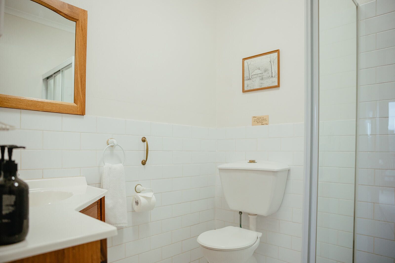 Et badeværelse med et hvidt håndvaskarrangement, toilet og bruseområde. Et spejl er monteret over håndvasken, og der hænger et billede i ramme på væggen.