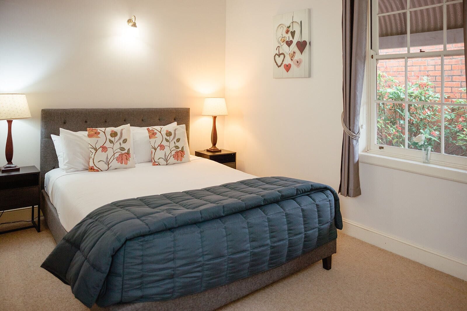 A bedroom with a bed, two bedside lamps, a nightstand, a window with curtains, and a wall decoration.