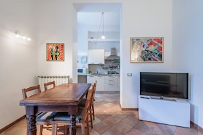 A dining area with a wooden table and chairs, a kitchen in the background, a TV on a stand, and two framed artworks on the walls.