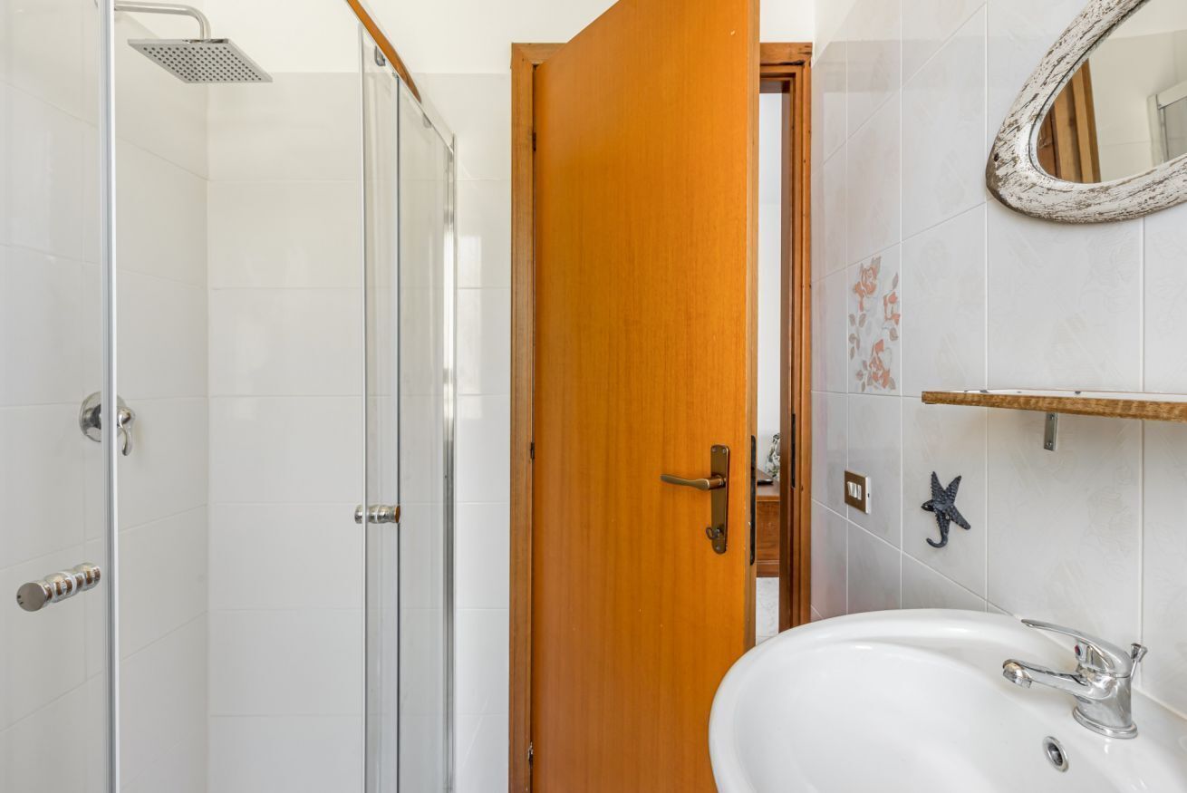 A bathroom with a glass shower enclosure, a wooden door, a white sink, and a mirror on the wall.