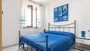 A room with a bed, a nightstand, a window with curtains, and a wall with a geometric pattern. - GuestHost - Carloforte Cozy Apartment with Balcony (Carloforte)