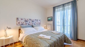 Un dormitorio con cama, mesita de noche, radiador y ventana con cortinas.