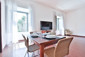 A dining area with a wooden table, wicker chairs, a flat-screen TV, and large windows with white curtains.