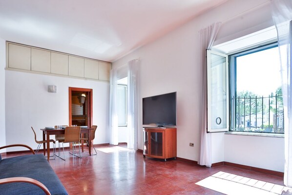 A room with a dining table, chairs, a TV, a sofa, and a large window with a view.