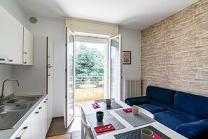 Un espacio habitable compacto con kitchenette, una zona de comedor con mesa y sillas, un sofá azul y vistas a un balcón con mobiliario exterior.