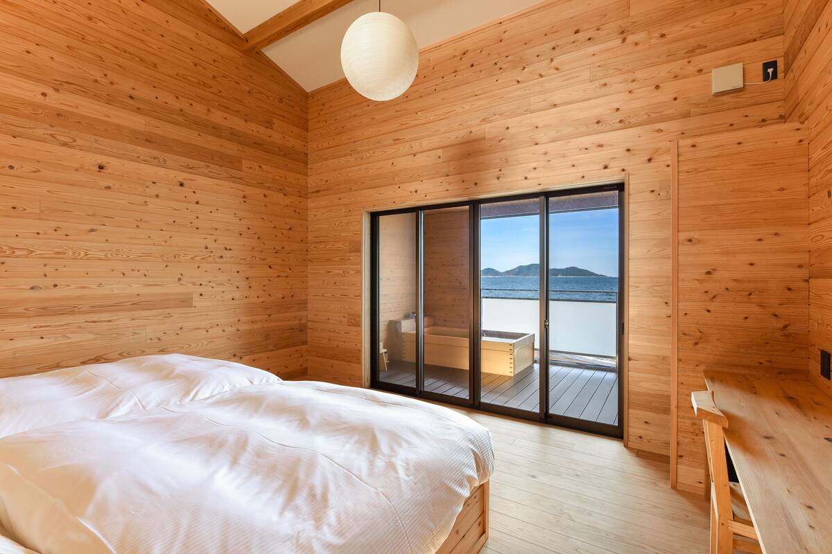 A wooden cabin bedroom with a bed, a wooden bench, and a sliding glass door leading to a balcony with a view of the sea.