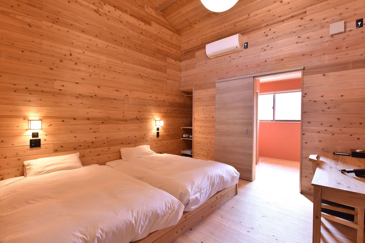 A wooden cabin room with two beds, a desk, and an air conditioning unit.