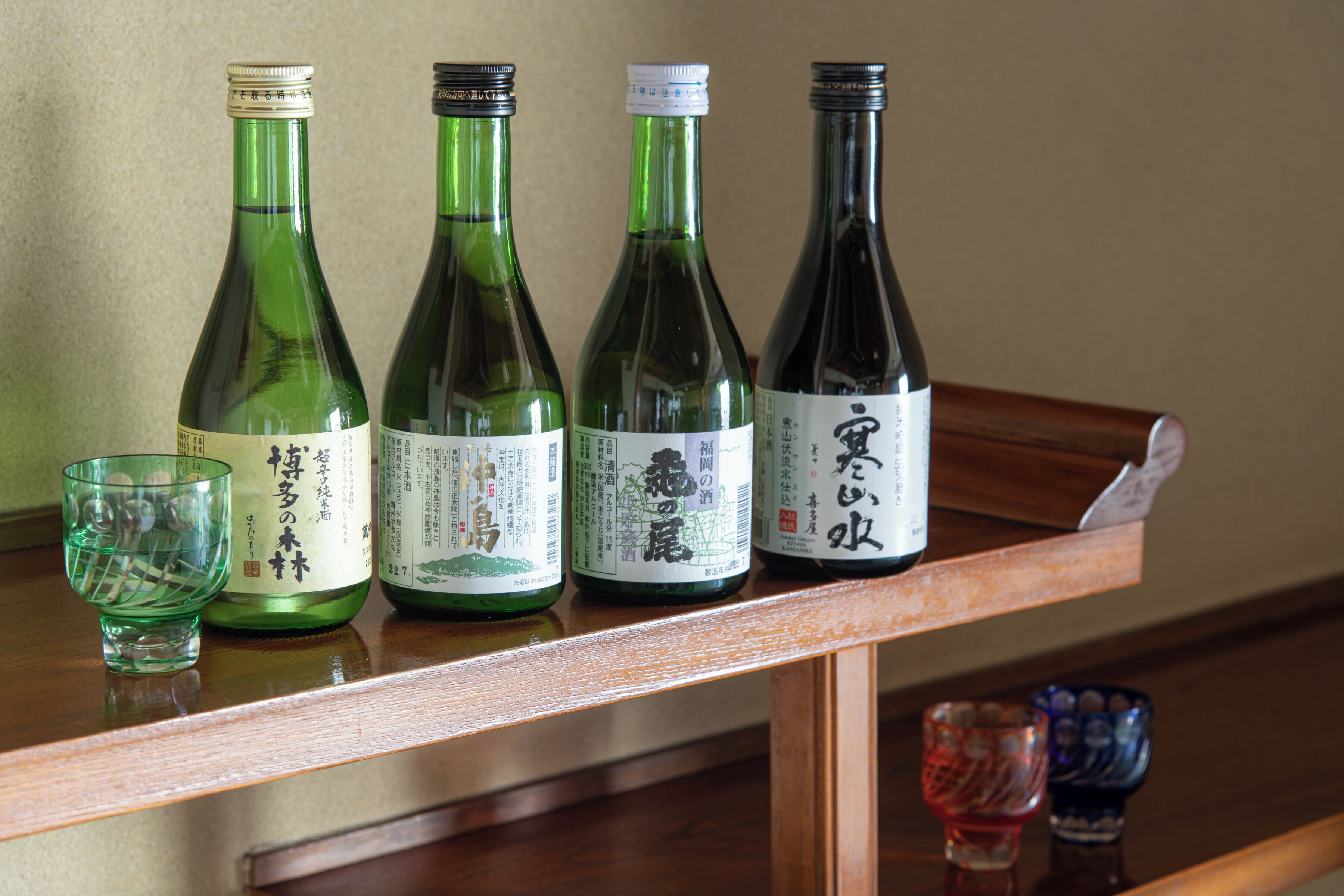 Vier flessen sake op een houten plank, met een glas en twee kleine bekertjes eronder.