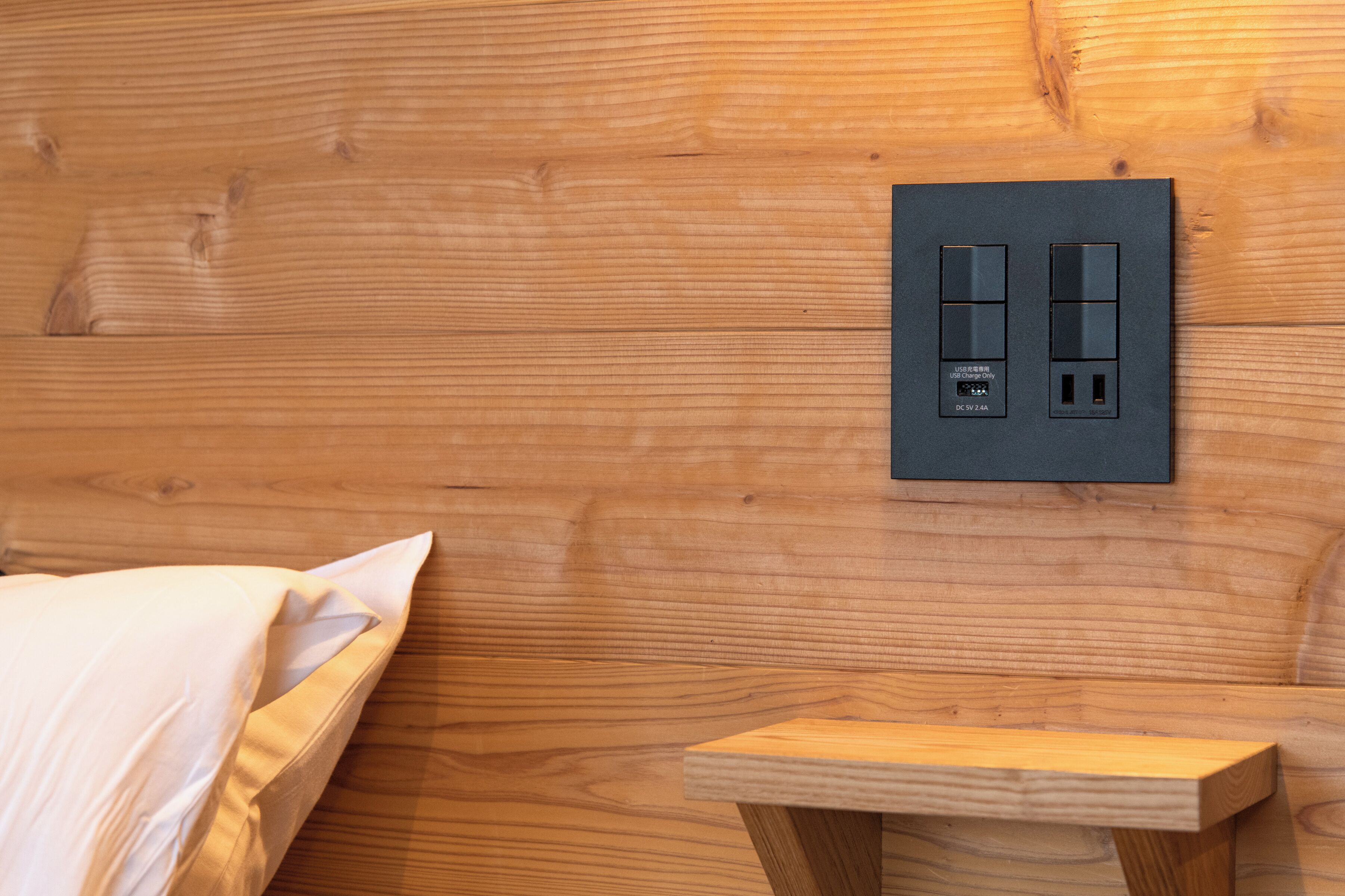 A wooden wall with a black electrical outlet and a small wooden nightstand.