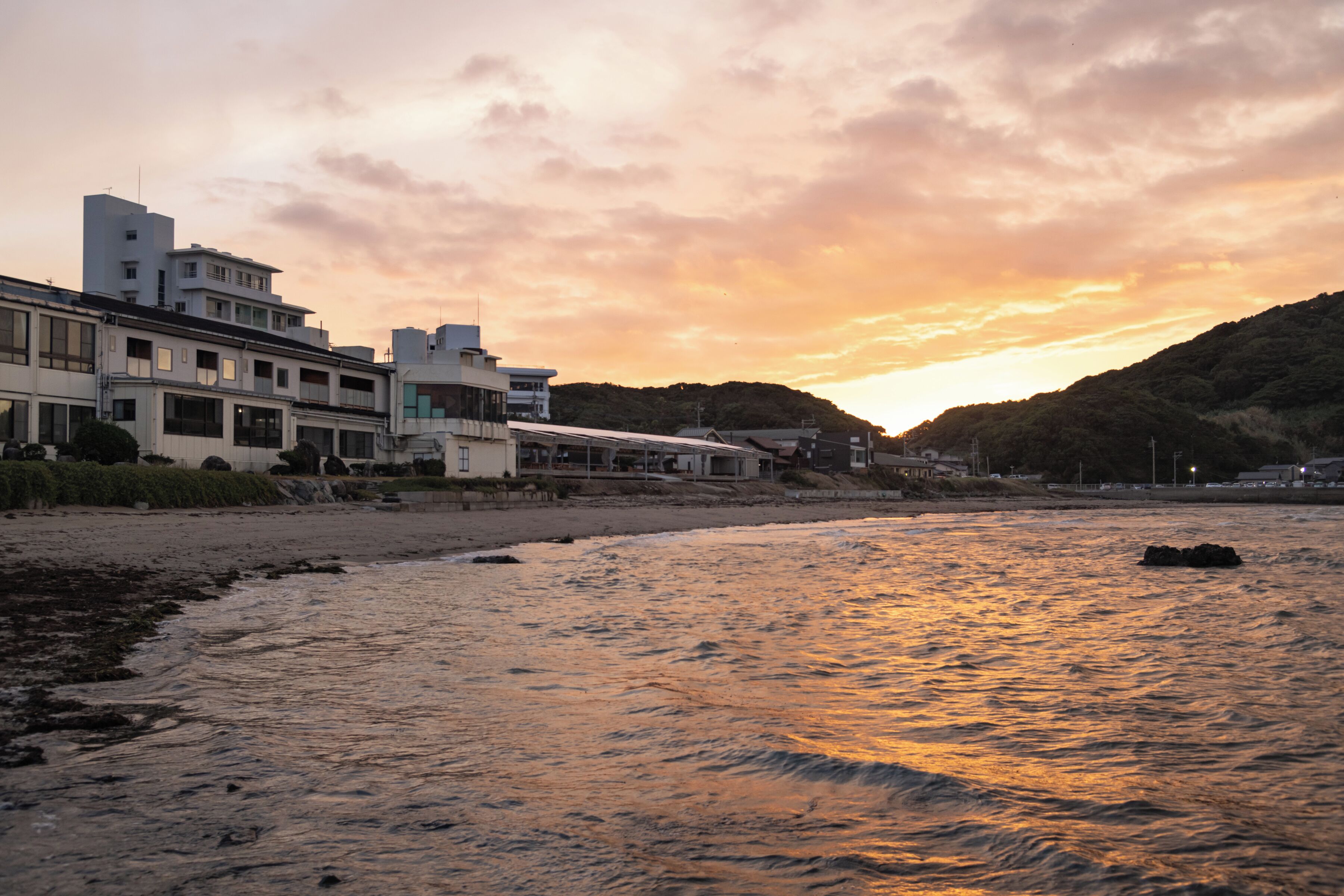 Un conjunto de edificios costeros con varias construcciones, una playa en primer plano y una puesta de sol al fondo.
