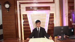 A man sitting at a wooden desk with a computer, writing in a notebook. - Hotel Rudreshwar (Haridwar)