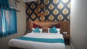 A neatly made bed with a wooden headboard, a nightstand with a phone and a lamp, and a patterned wallpaper on the wall. - Hotel Rudreshwar (Haridwar)