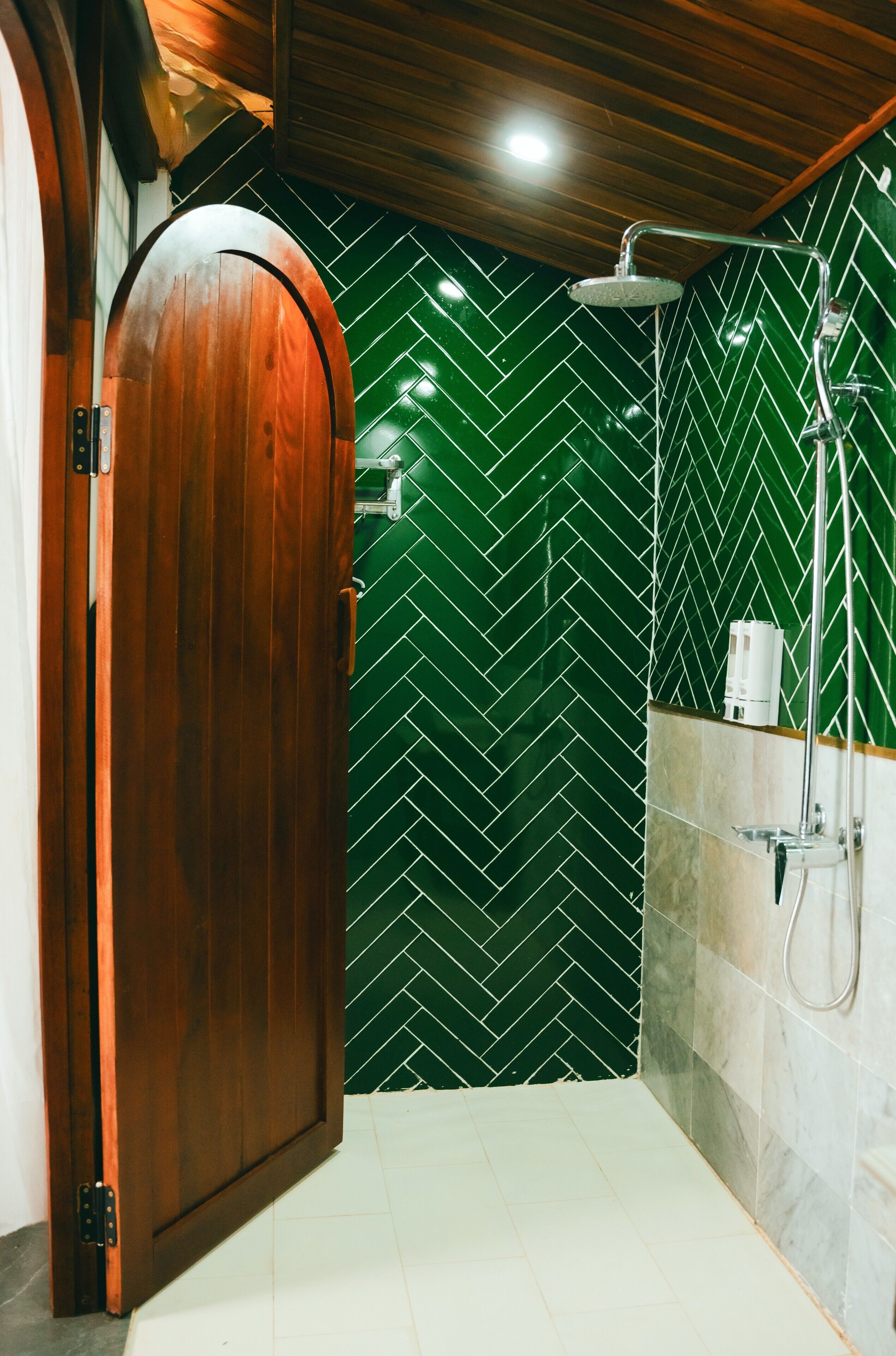 A bathroom with a wooden door, green herringbone patterned tiles, a shower area, and a marble wall.