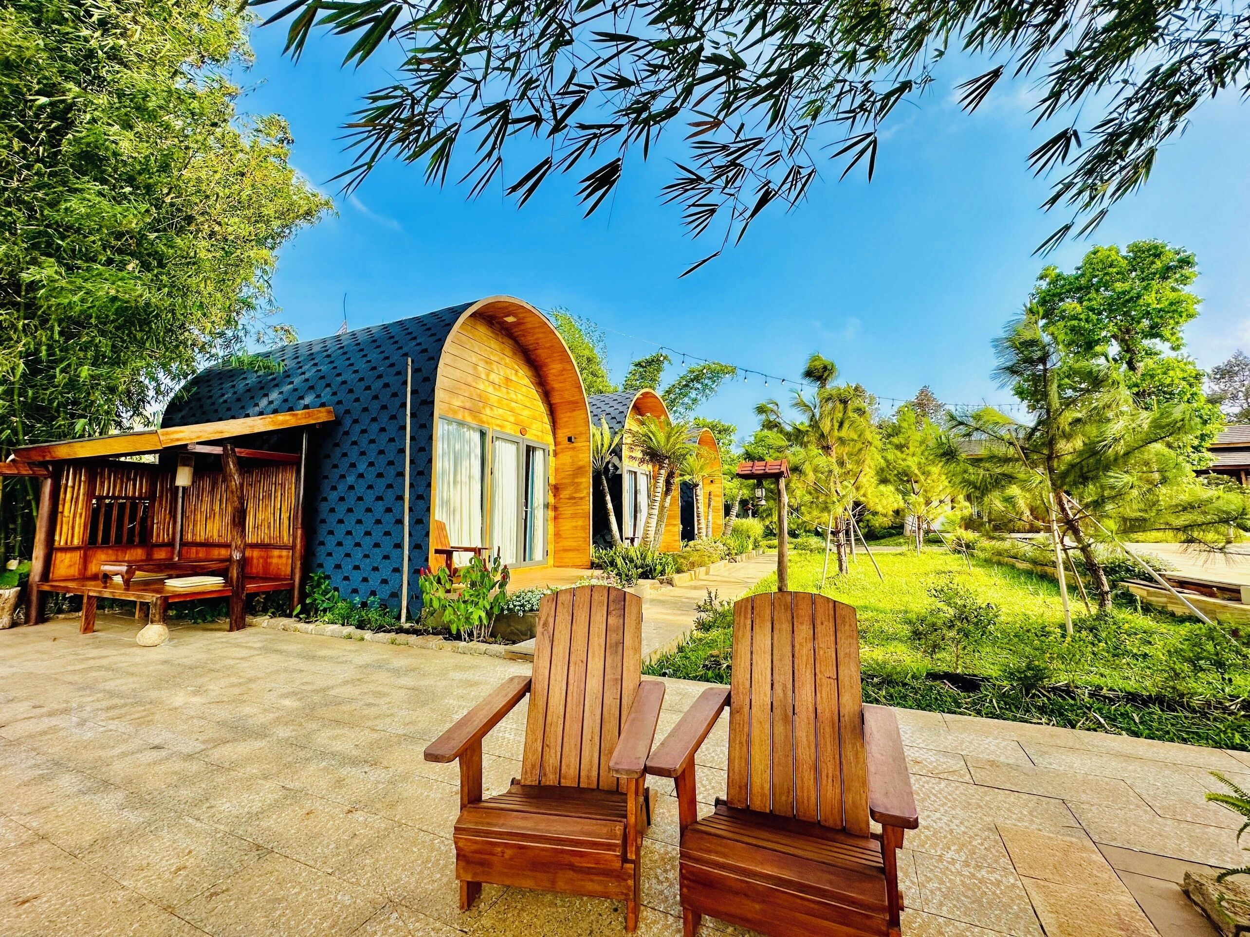 A wooden structure with a curved roof and large windows, surrounded by lush greenery and a clear blue sky.