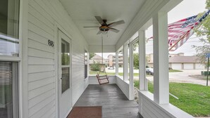 A covered porch with a ceiling fan, a swing, and a view of a residential area with an American flag. - New Kitchen,FirePit,FamFriendly,Sleeps10:LakeLux (Arnolds Park)