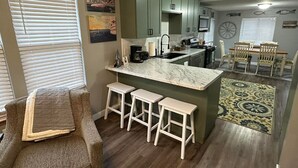 A compact kitchen with a central island, bar stools, and a dining area with a table and chairs. - New Kitchen,FirePit,FamFriendly,Sleeps10:LakeLux (Arnolds Park)