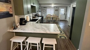 A kitchen with a marble countertop, white bar stools, and a dining area with a table and chairs. - New Kitchen,FirePit,FamFriendly,Sleeps10:LakeLux (Arnolds Park)