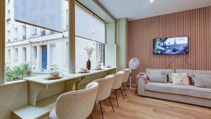 A modern living room with a sofa, a dining area with chairs and a table set, a TV mounted on the wall, and a view of the city through large windows.