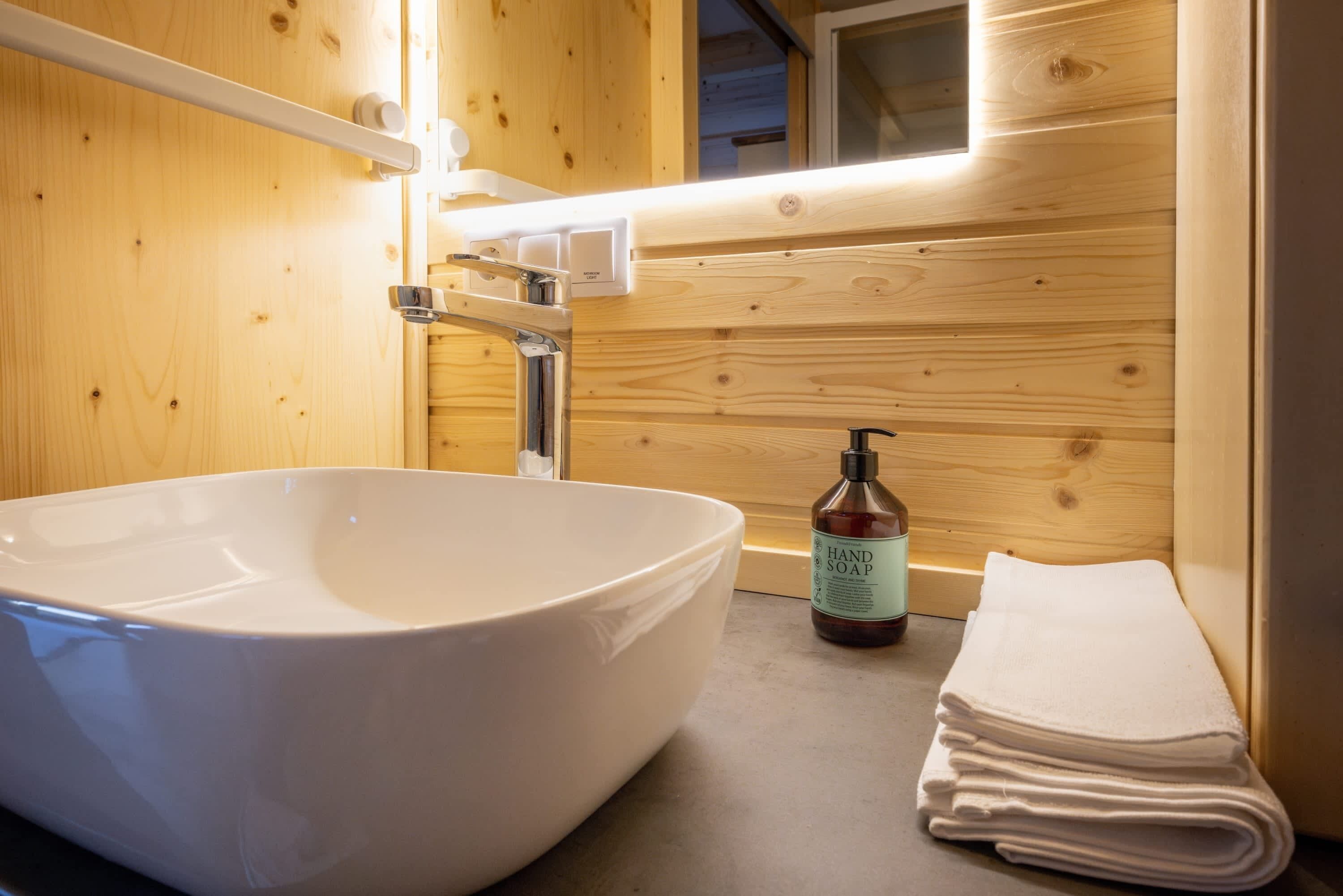 A bathroom with a large white sink, wooden walls, a mirror, a hand soap bottle, and a stack of towels.