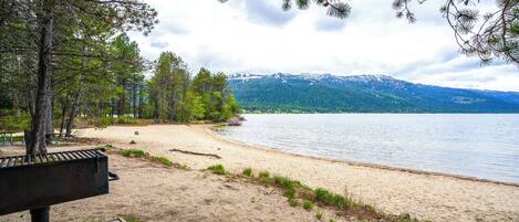 Blick auf einen See mit Sandstrand, Bäumen und Bergen im Hintergrund.