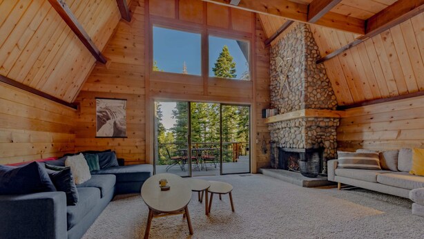 L’intérieur chaleureux d’un chalet en bois, avec une cheminée, une grande fenêtre donnant sur les arbres et un coin salon confortable.