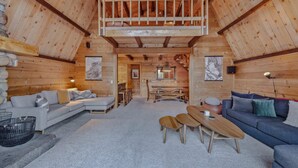 L’intérieur d’une cabane en rondins comprend une chambre à coucher située en mezzanine, un espace de vie avec un canapé et une table basse, ainsi qu’un coin repas avec des bancs.