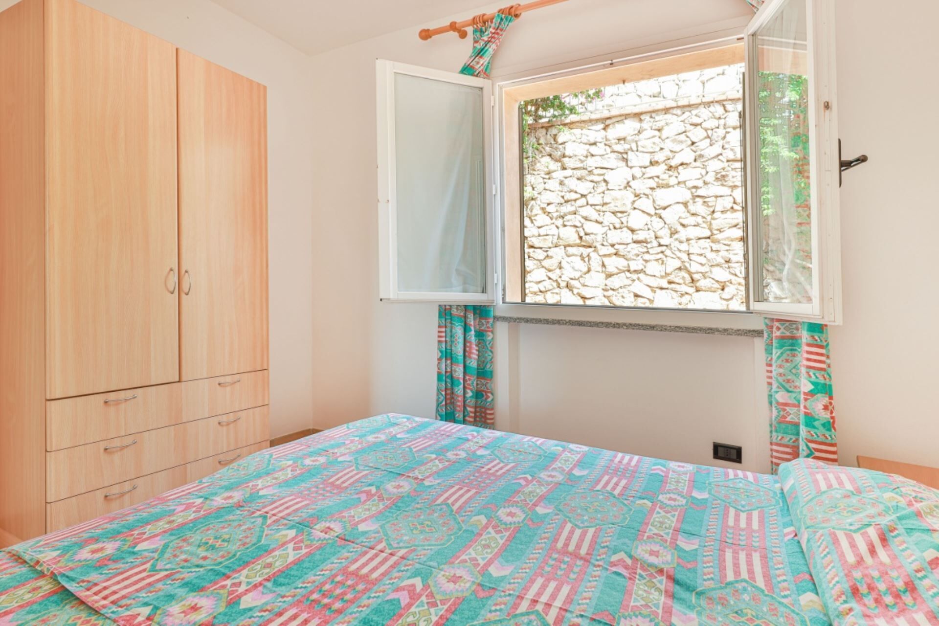 A bedroom with a bed, a wardrobe, a window with a view of a stone wall, and curtains.