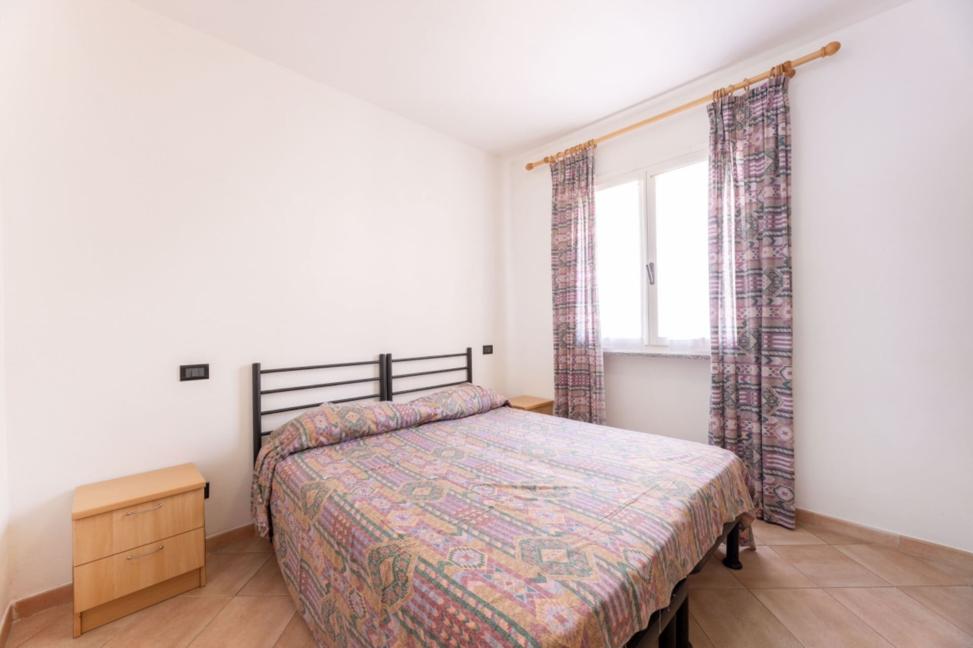 Een slaapkamer met een bed, een houten nachtkastje, een raam met gordijnen met een patroon, en een effen witte muur.