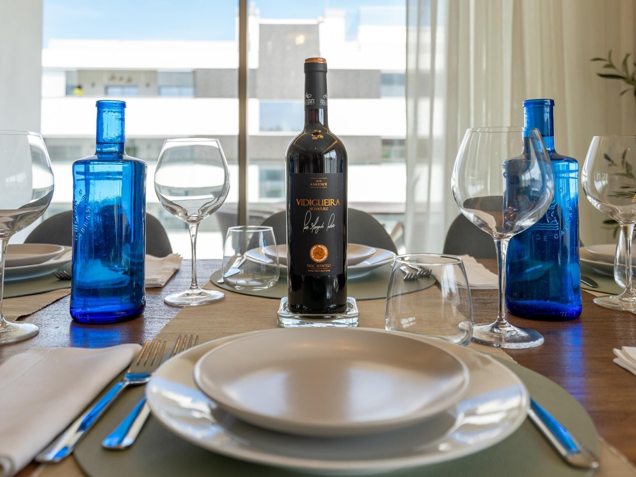 Mesa de jantar posta com pratos, talheres e copos de vinho, com uma garrafa de vinho em destaque.