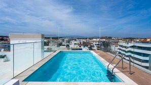 Une piscine sur le toit, avec une eau d’un bleu cristallin, entourée de rampes de verre et d’une terrasse en bois.