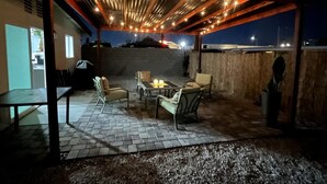 Un patio al aire libre con una pérgola adornada con luces de cuerda, un brasero y mobiliario exterior.