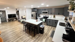 Una cocina moderna con armarios grises, encimera blanca y electrodomésticos de acero inoxidable. Hay un comedor con mesa y sillas, y una pequeña zona de estar con sofá.