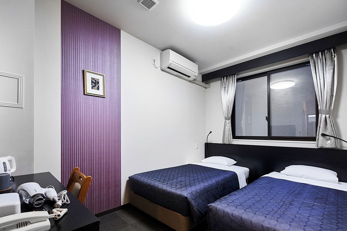 A hotel room with two beds, a desk with a phone and a hairdryer, a window with curtains, and an air conditioning unit.