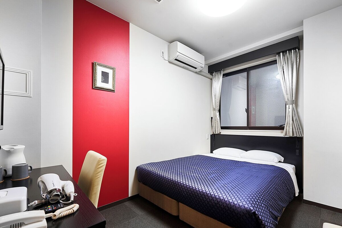 A hotel room with a bed, a desk with a telephone, a chair, an air conditioning unit, and a window with curtains.
