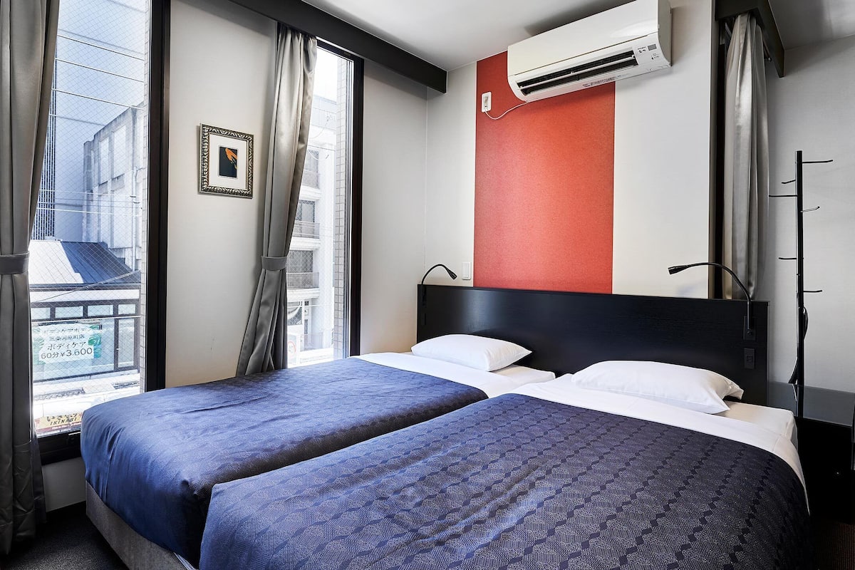 A hotel room with two beds, a window with curtains, an air conditioning unit, and a view of the outside.