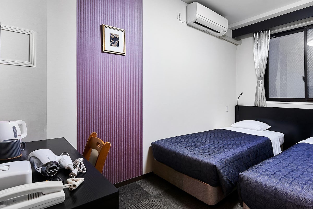 A hotel room with two beds, a desk with a phone and a hairdryer, a purple wall, and a window with curtains.
