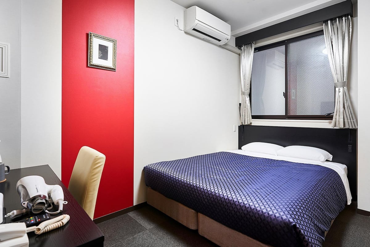 A hotel room with a red accent wall, a bed with a blue patterned bedspread, a desk with a hairdryer, and a chair.