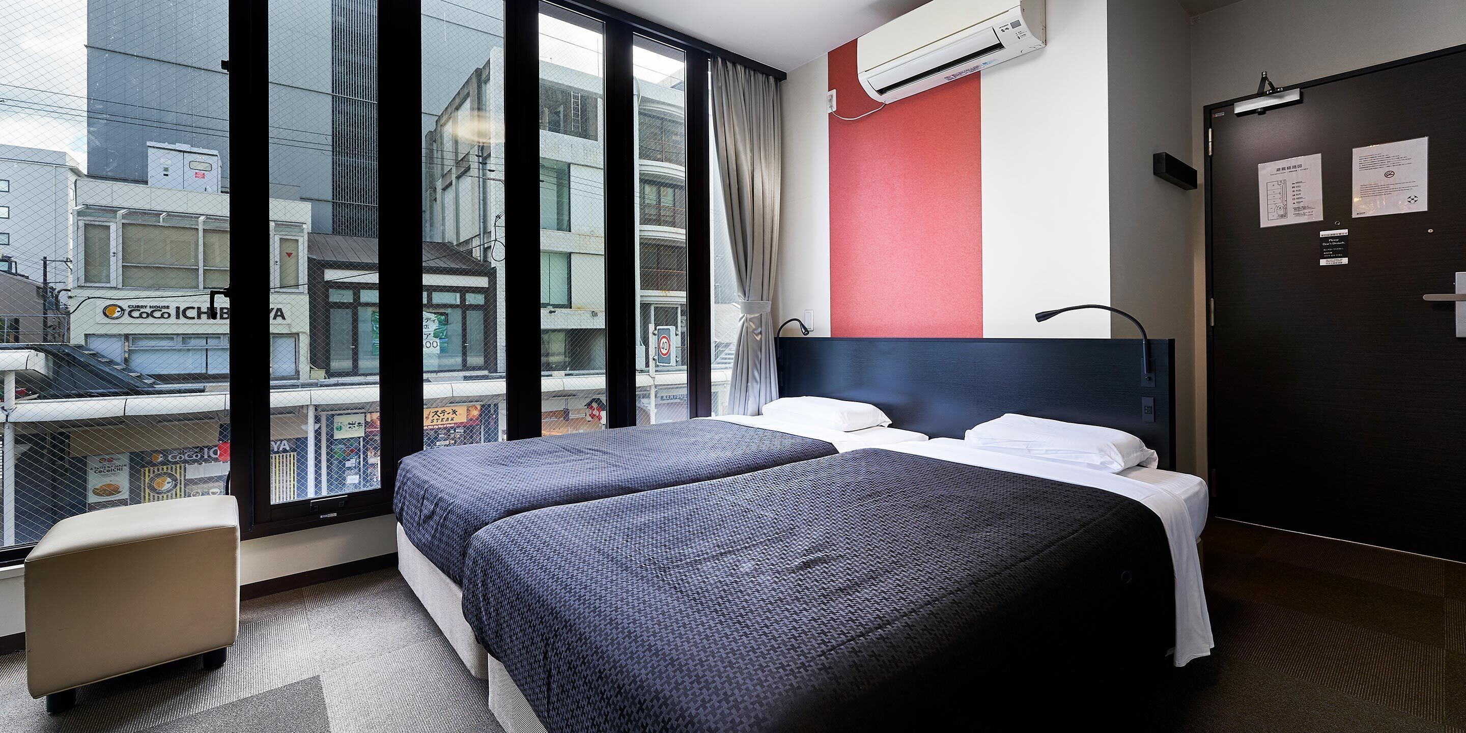 A hotel room with two beds, a window with a view of the city, and an air conditioning unit on the wall.