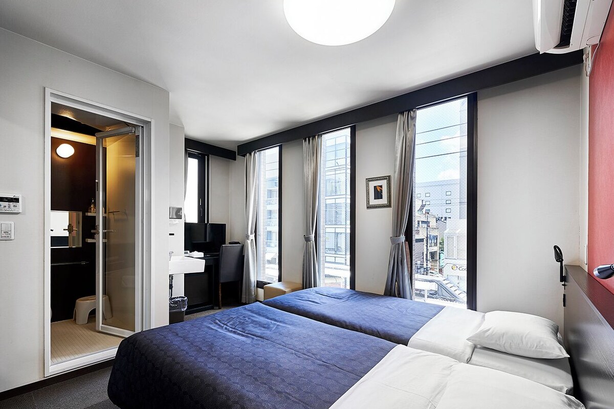 A hotel room with two beds, a desk, a chair, a bathroom, and a view of the city.