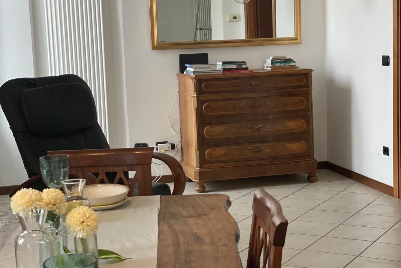 A dining area with a wooden table, chairs, a wooden dresser with books, a mirror, and a vase with flowers.