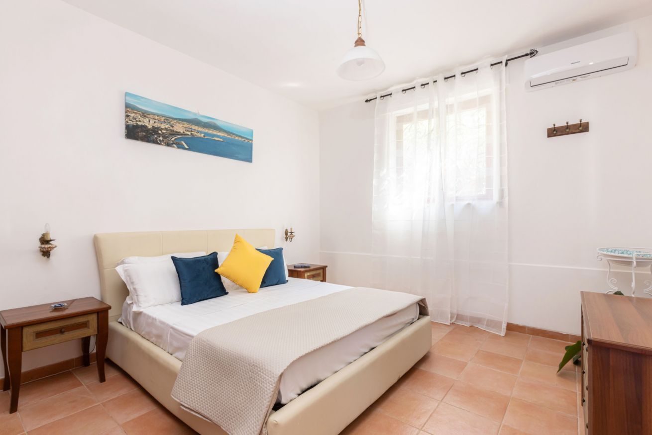 A bedroom with a bed, bedside tables, a nightstand, a window with curtains, a wall-mounted air conditioner, and a painting on the wall.