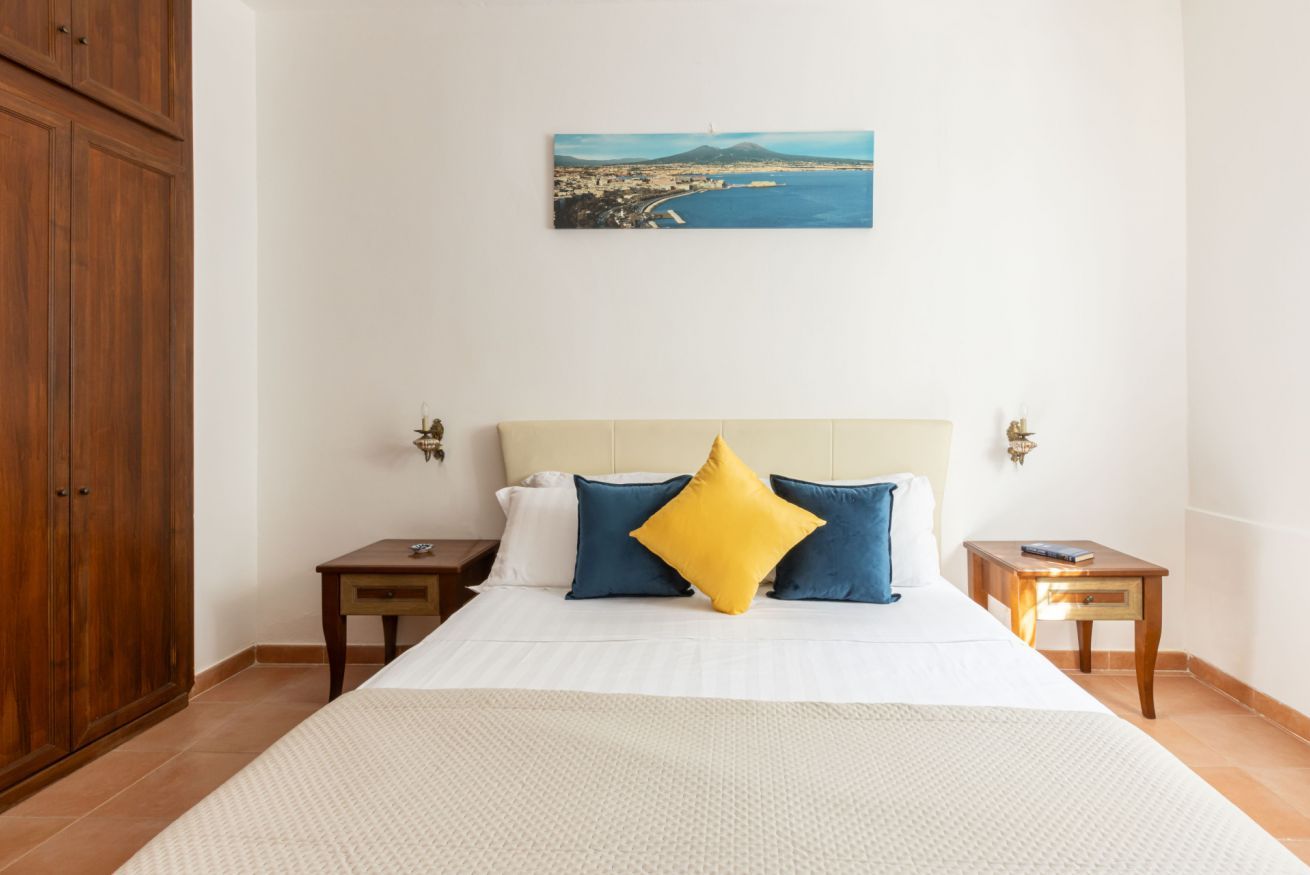 A bedroom with a bed, two nightstands, a wooden wardrobe, and a painting of a coastal landscape.