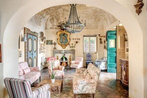 Una stanza con soffitto a volta alto, un lampadario e un camino. Sono presenti diverse poltrone imbottite e una poltrona a righe.