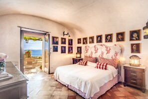 Une chambre avec un grand lit, une vue sur la mer par une porte ouverte et un mur décoré de tableaux encadrés.