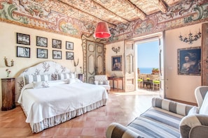 A spacious bedroom with a large bed, a chair, and a view of the sea through an open door.