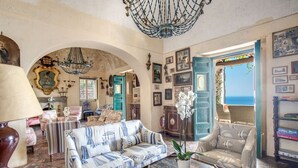 A spacious living room with a large sofa, armchairs, and a wooden coffee table. There is a chandelier, framed artwork, and a view of the sea through an open door. - MyExclusiveItaly - Lux Historic Villa with Pool & Amalfi Coast View (Positano)