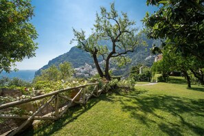 Un giardino pittoresco con un gazebo in legno, vegetazione lussureggiante e vista su una cittadina costiera e montagne sullo sfondo.
