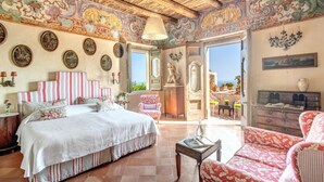 A spacious bedroom with a large bed, a chair, and a desk. There are multiple framed pictures on the walls and a chandelier. - MyExclusiveItaly - Lux Historic Villa with Pool & Amalfi Coast View (Positano)